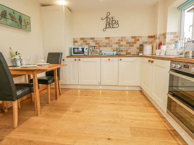 A kitchen with white cabinets wooden flooring a dining table with black chairs and kitchen appliances at Snowdrop Cottage in Aldingham