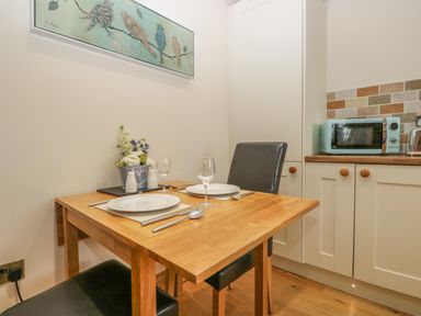 A kitchen dining area with a wooden table set for two with plates and glasses next to a cabinet with a microwave and toaster at Snowdrop Cottage in Aldingham