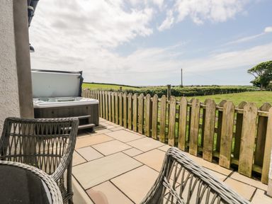An outdoor patio with wicker chairs a hot tub and a wooden fence overlooking fields at Snowdrop Cottage in Aldingham