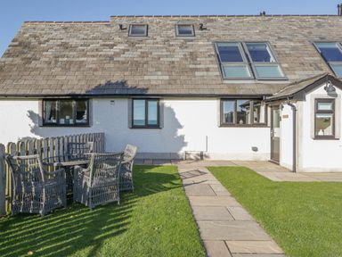 A house exterior with a stone roof a paved pathway a grassy yard and wicker chairs and a table on the grass at Bluebell Cottage in Aldingham