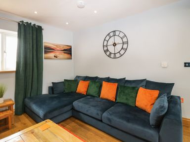A living room with a large blue sectional sofa with green and orange cushions a wall clock and a window with green curtains at Bluebell Cottage in Aldingham