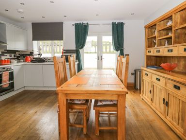 A kitchen with wooden dining table and chairs next to wooden cupboard and white cabinets at Bluebell Cottage in Aldingham