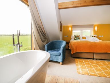 A bedroom with an orange bedspread a blue armchair a freestanding bathtub and a large window at Bluebell Cottage in Aldingham