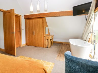 A bedroom with wooden doors a wardrobe a desk with stools a wall-mounted tv a freestanding bathtub and a blue chair at Bluebell Cottage in Aldingham
