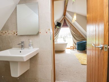 A bathroom sink with mirror beside a room with a bathtub blue chair and window with curtains at Bluebell Cottage in Aldingham