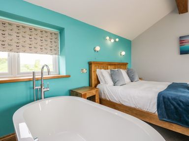 A bedroom with a wooden bed white bedding and gray pillows a bathtub near a window with butterfly patterned blinds at Bluebell Cottage in Aldingham
