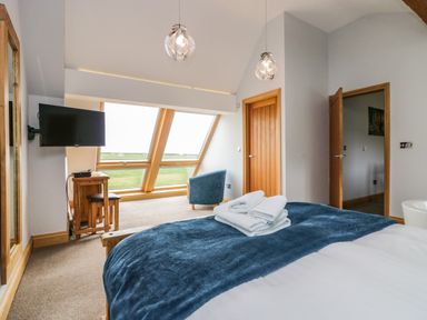 A bedroom with a bed folded towels a wall mounted tv a wooden desk with stools a blue chair and large angled windows at Bluebell Cottage in Aldingham