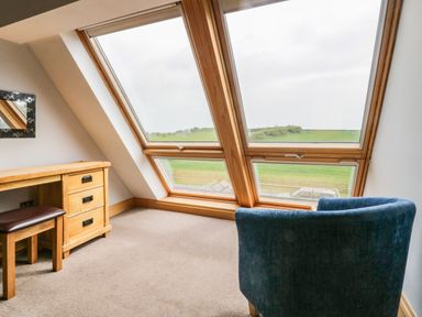 A room with large slanted windows a wooden desk a stool and a blue armchair at Bluebell Cottage in Aldingham