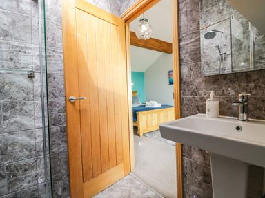 A bathroom with a shower, sink, and open wooden door leading to a bedroom with a bed and folded towels at Bluebell Cottage in Aldingham