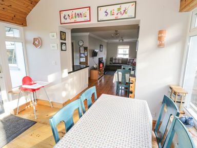 A dining area with a table covered by a patterned tablecloth surrounded by blue chairs next to a window and a red high chair near the door at Teach Teolai in Carraroe County Galway