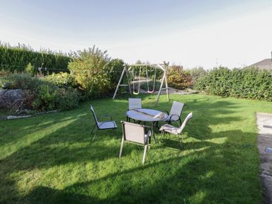 A garden with a glass table and five chairs and a swing set surrounded by bushes at Teach Teolai in Carraroe County Galway