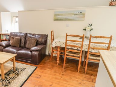 A room with a brown leather sofa with cushions a wooden dining table with chairs and a flower vase on the table at Hendoll Cottage 2 Fairbourne near Dolgellau