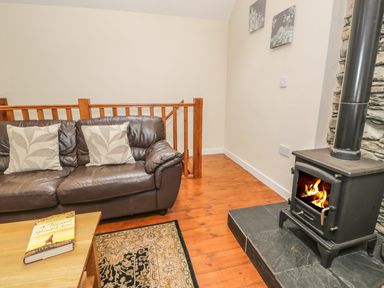 A living room with a leather sofa two pillows a coffee table with a book a rug and a wood burning stove at Hendoll Cottage 2 in Fairbourne near Dolgellau