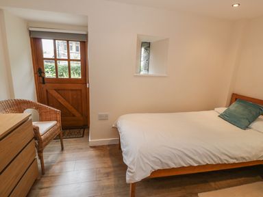 A bedroom with a wooden bed and white bedding a wicker chair a wooden dresser and a wooden door at Hendoll Cottage 2 Fairbourne near Dolgellau