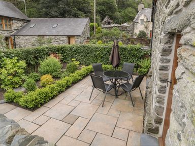 A stone patio with a round glass table and four chairs with an umbrella in a garden surrounded by stone buildings at Hendoll Cottage 2 Fairbourne near Dolgellau