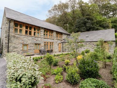 A stone house with wooden windows and doors next to a garden with plants and shrubs at Hendoll Barn Friog near Fairbourne