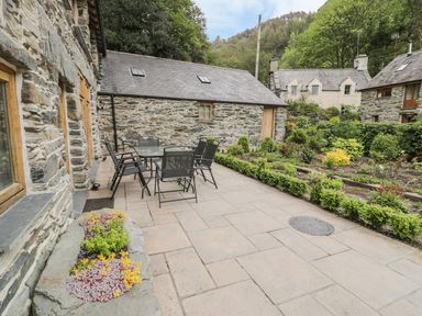 A stone patio with a glass table and six chairs next to a garden and stone buildings at Hendoll Barn Friog near Fairbourne