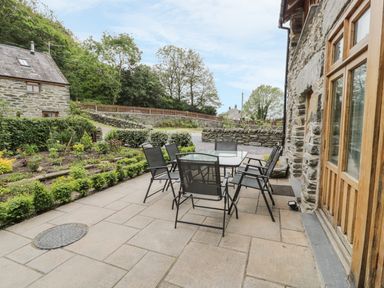 An outdoor patio with a glass table and six chairs next to a stone house at Hendoll Barn in Friog near Fairbourne