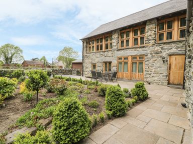 A stone house with wooden windows and doors next to a garden and patio with table and chairs at Hendoll Barn Friog near Fairbourne
