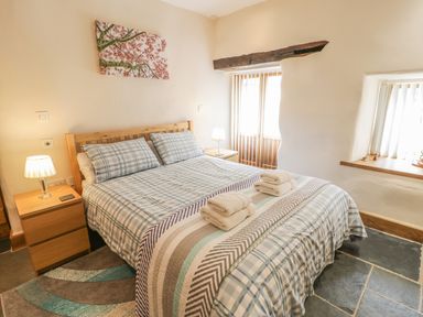 A bedroom with a double bed with plaid bedding two bedside tables with lamps a wall painting and a window with wooden beam at Hendoll Barn in Friog near Fairbourne