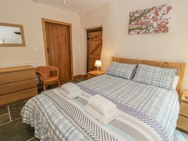 A bedroom with a double bed with checked bedding towels on the bed wooden doors a wicker chair a dresser and a lamp at Hendoll Barn in Friog near Fairbourne