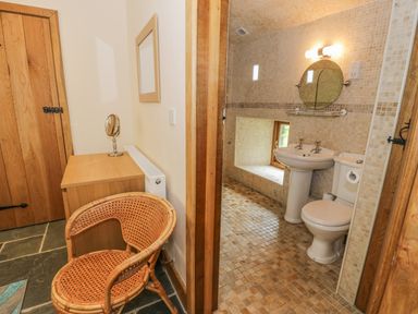A bathroom with a toilet sink round mirror and a wicker chair next to a wooden cabinet at Hendoll Barn Friog near Fairbourne