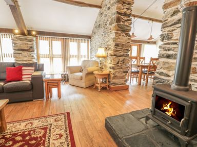 A living room with a wood stove a gray sofa and an armchair next to stone pillars and a dining table in the background at Hendoll Barn in Friog near Fairbourne
