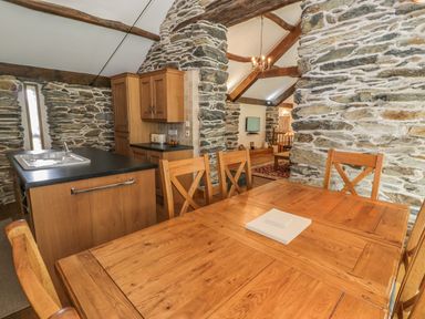 A dining table with chairs and a kitchen area with wooden cabinets and stone walls at Hendoll Barn in Friog near Fairbourne