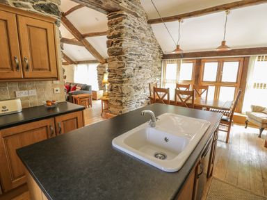 A kitchen island with a sink overlooks a dining area with a wooden table and chairs and a living room with sofas and wooden beams at Hendoll Barn Friog near Fairbourne