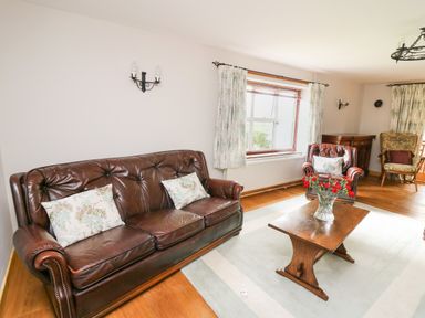 A living room with a leather sofa and armchair a wooden coffee table with flowers and floral curtains at Holly Farm Cottage in Howey