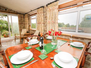 A dining area with a wooden table set for six with white dishes a wine bottle and a vase with red flowers next to windows and two armchairs at Holly Farm Cottage in Howey