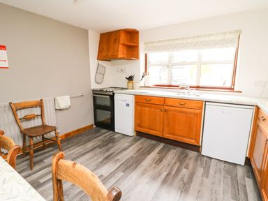 A kitchen with wooden cabinets a black oven a white dishwasher a small white fridge and wooden chairs at Holly Farm Cottage in Howey