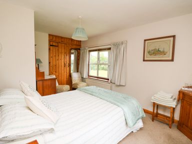 A bedroom with a double bed, wooden wardrobe, two chairs, a window with curtains, framed wall art, and folded towels on a wooden stool at Holly Farm Cottage in Howey