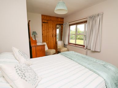 A bedroom with a bed, two chairs, a wooden wardrobe, a desk with a lamp, and a window with curtains at Holly Farm Cottage in Howey