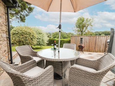 An outdoor patio with a round glass table and four woven chairs under an umbrella with a bottle of wine and glasses at Holly Farm Cottage in Howey
