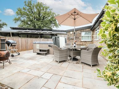 An outdoor patio with wicker chairs around a glass table under a large umbrella a hot tub a barbecue grill and a chiminea at Holly Farm Cottage in Howey