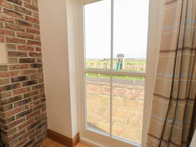 A window with a brick wall to the left and curtain to the right looking out to a stone patio and a children's playset at Grey Croft in Embleton