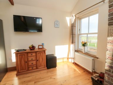 A room with a wall-mounted TV a wooden cabinet with fruit and decor a window with a vase of flowers and a radiator at Grey Croft in Embleton
