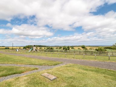 A playground with a slide and climbing frame surrounded by grass and a gravel path at Grey Croft in Embleton