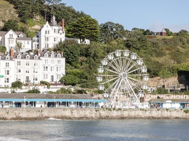 A waterfront view with white buildings a ferris wheel and a hillside with trees and a small structure at Treetops in Rhos-On-Sea