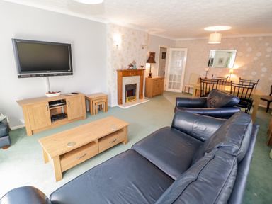 A living room with black leather sofas a wooden coffee table a wooden TV stand flat screen TV fireplace and a dining table with chairs at Treetops in Rhos-On-Sea