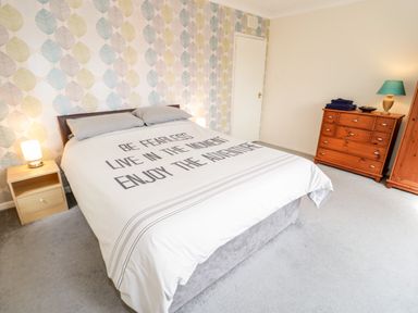 A bedroom with a bed with a printed duvet cover a wooden nightstand with lamp a wooden chest of drawers with folded towels and a lamp and a wallpapered wall at Treetops in Rhos-On-Sea
