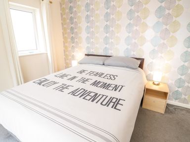 A bedroom with a bed featuring bedding with text a wooden nightstand with a lamp and leaf patterned wallpaper at Treetops in Rhos On Sea