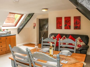 A dining area with a wooden table and chairs set for four with a bottle of wine and a black leather sofa with red and floral cushions in a room with a skylight window and wooden cabinets at Fig Trees - Wibble Farm in Williton near Watchet