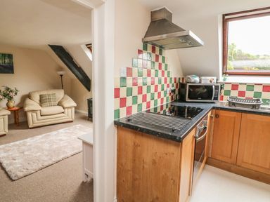 A kitchen with wooden cabinets and tiled backsplash next to a living room with cream armchairs and a carpet at Fig Trees - Wibble Farm in Williton near Watchet