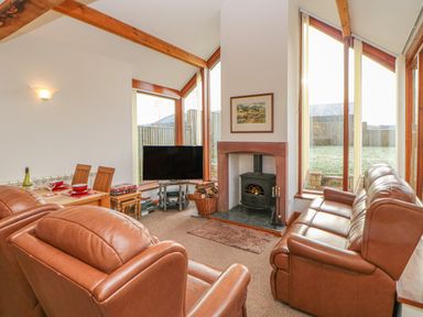 A living room with leather sofas a wood-burning stove a television wooden dining table and large windows at Swallow Cottage Milton near Brampton