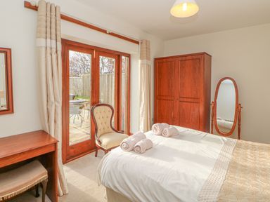 A bedroom with a bed towels a chair wooden wardrobe full length mirror desk and glass doors leading outside at Swallow Cottage in Milton near Brampton