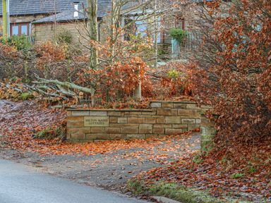 A stone wall with a sign saying Milton Mains Cottages surrounded by trees with autumn leaves on the ground at Swallow Cottage in Milton near Brampton