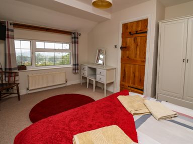 A bedroom with a bed covered with towels and a red blanket a wooden door a white wardrobe a white dressing table with a mirror a red round rug and a window with curtains at Aberdauddwr in Lampeter