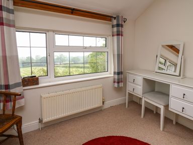 A room with a window overlooking fields a white dressing table with mirror a cushioned stool a wooden chair and patterned curtains at Aberdauddwr in Lampeter
